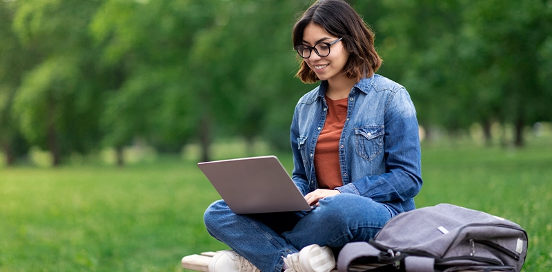 Online student sitting on a park bench studying on her laptop computer.